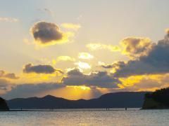 海の遠方に見える島と雲の切れ間から太陽のオレンジ色の光が差し込んでいる写真