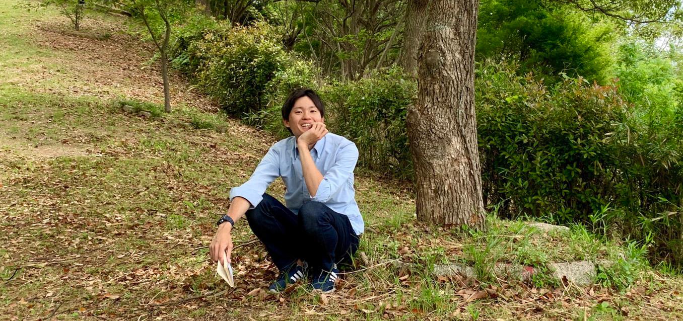 田山 直樹さんが木の前でしゃがんで座っており左手で顔を支えて笑顔で写っている写真