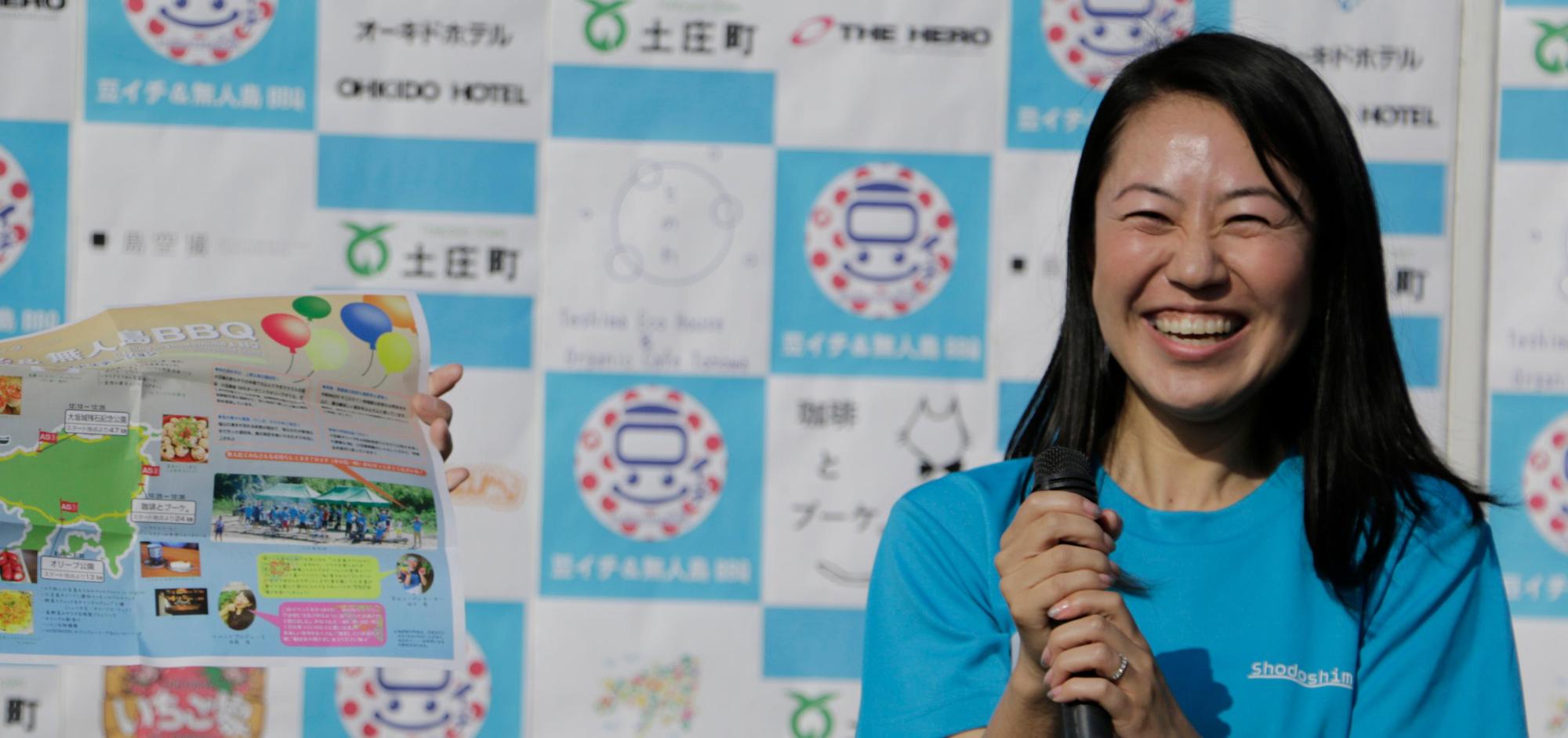 青いティシャツを着て、両手でマイクを持ち満面の笑みで写っている須藤 渚さんの写真